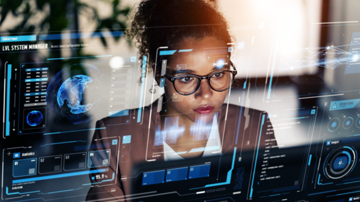 A person with a blurred face is working on a futuristic, transparent computer interface. The interface displays various digital elements, including graphs, charts, and data visualizations.