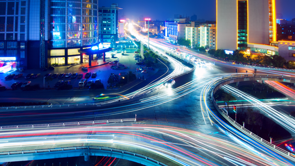 Nighttime cityscape with highways and overpasses, featuring long exposure light trails of vehicles and tall buildings with illuminated windows and signs.