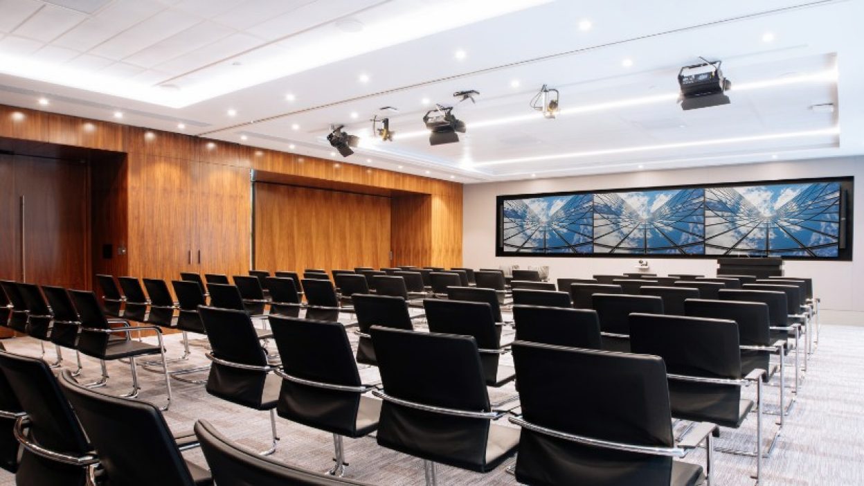 Forum set theatre style with black chairs and silver arms and legs. Wooden interiror, fixed ceiling lighting and three large screens at the front of room with the Sky atrium image on all three.