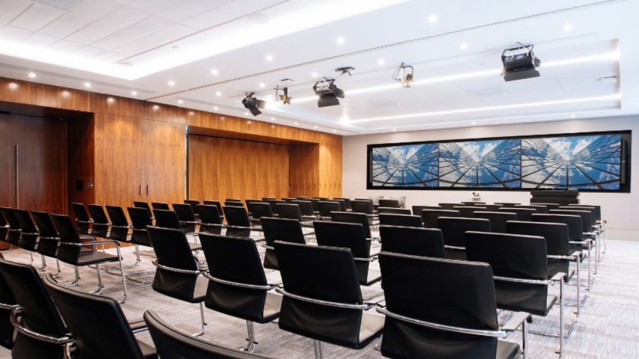 Forum set theatre style with black chairs and silver arms and legs. Wooden interiror, fixed ceiling lighting and three large screens at the front of room with the Sky atrium image on all three.