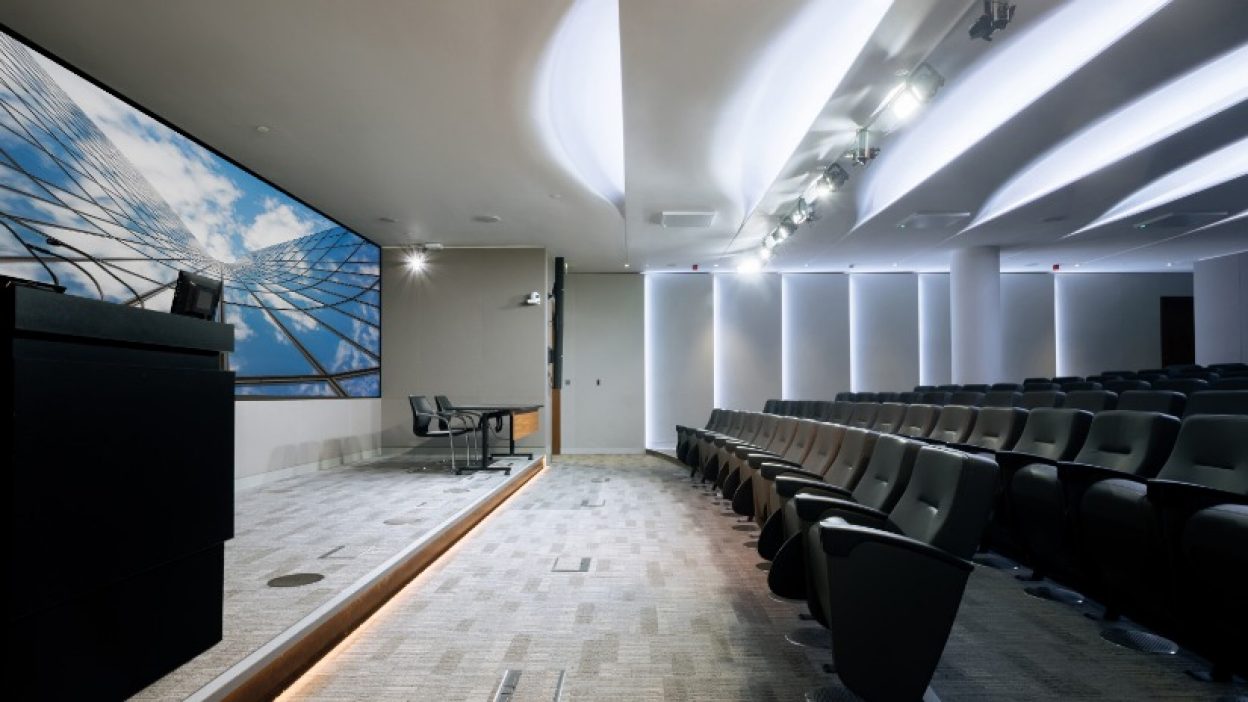 Tiered auditorium side shot with lectern and top table. Sky atrium image on the video wall.